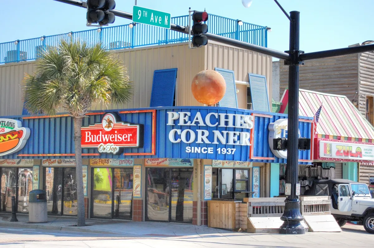 A lively street scene with the iconic Peaches Corner sign, colorful awnings, palm trees, and people enjoying drinks and dining. The image exudes an energetic nightlife atmosphere and highlights the proximity to local cuisine, social venues, and sunset views.