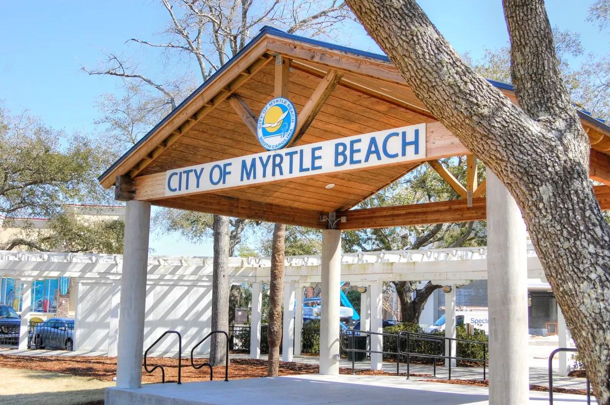The image captures a welcoming wooden structure with signage, surrounded by trees. The scene suggests local community events, gatherings, and access to information about happenings in Myrtle Beach, reinforcing the active, social aspect of a long-term winter stay.
