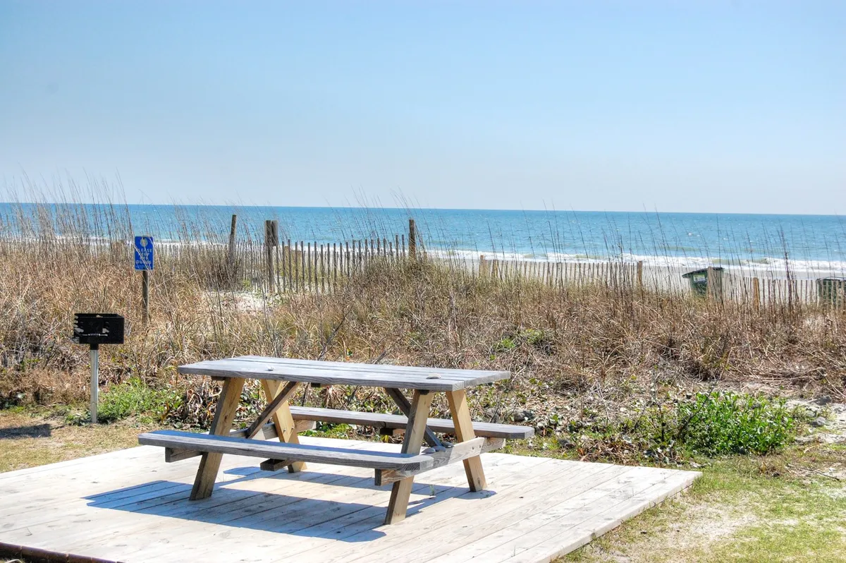 A serene picnic table surrounded by dune grass, with an open view to the ocean and a fence line in the distance. This peaceful setting is perfect for sunset gatherings, morning coffee, or simply unwinding with friends and loved ones by the sea.