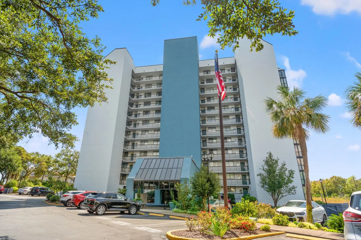 This image shows the facade of Seamark Towers, featuring the entrance awning, a welcoming flag pole, palm trees, and the parking area. The sunlit exterior and inviting surroundings set a positive first impression, hinting at the welcoming community snowbirds can expect during their stay.