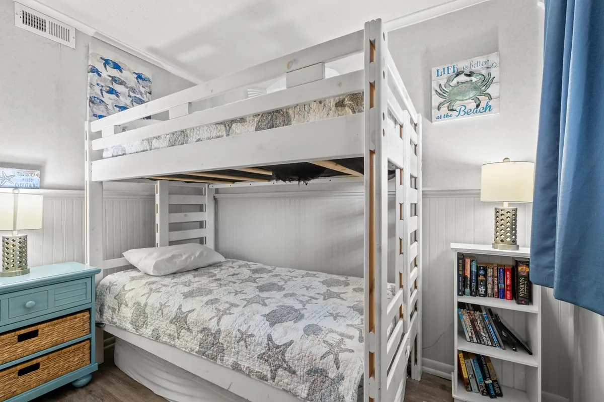 A cheerful twin bunk room with a colorful quilted bunk bed, a nightstand with a lamp, and a bookshelf full of books, creating a playful yet restful environment for kids or friends.