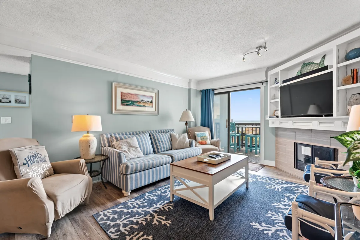 The image shows a cozy, beautifully furnished living room with a striped sofa and coffee table, centered around a welcoming fireplace. Large windows flood the space with natural light and showcase stunning views of the ocean, inviting guests to relax and soak in the scenery.