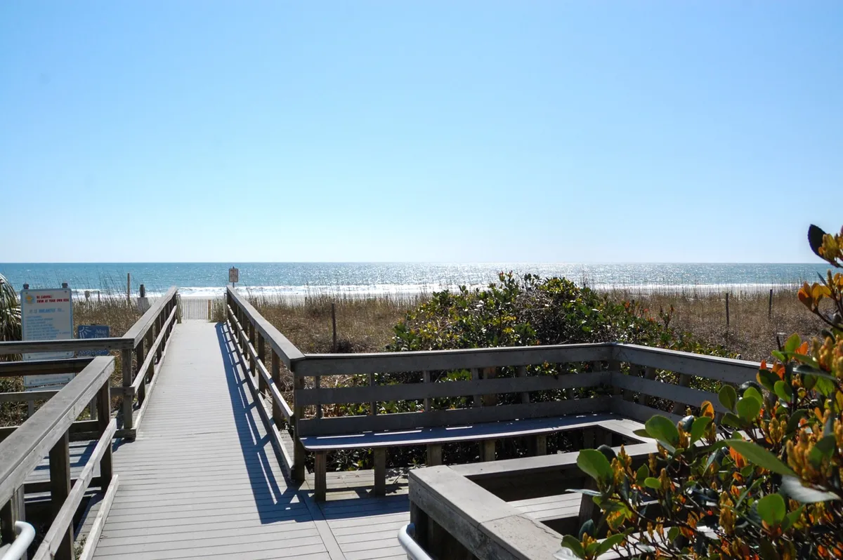 A scenic boardwalk leads through dune vegetation directly to the beach, with the ocean visible in the background. The inviting path promises effortless access to sand and surf, reinforcing the beachside lifestyle.