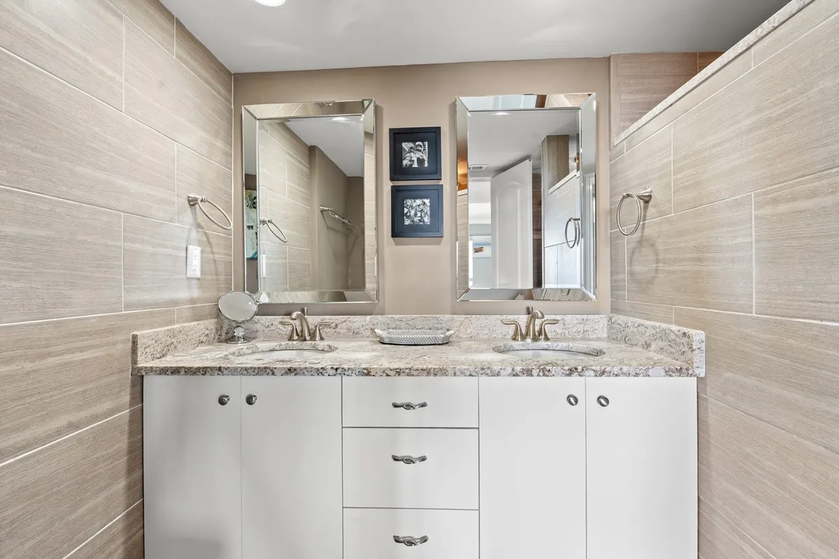 A modern bathroom featuring a double vanity with a granite countertop, two mirrors, and calming wall art. The space feels fresh, well-lit, and spacious, perfect for accommodating busy mornings or winding down after the beach.