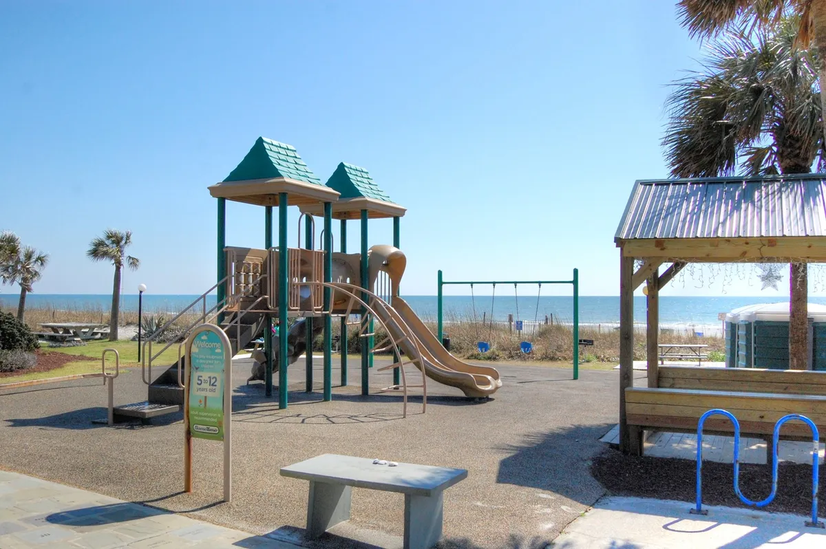 A vibrant playground featuring swings and climbing equipment with the sandy beach and palm trees in the background. Children are playing and families are enjoying relaxed picnics nearby, set against a sunny, inviting oceanfront scene.