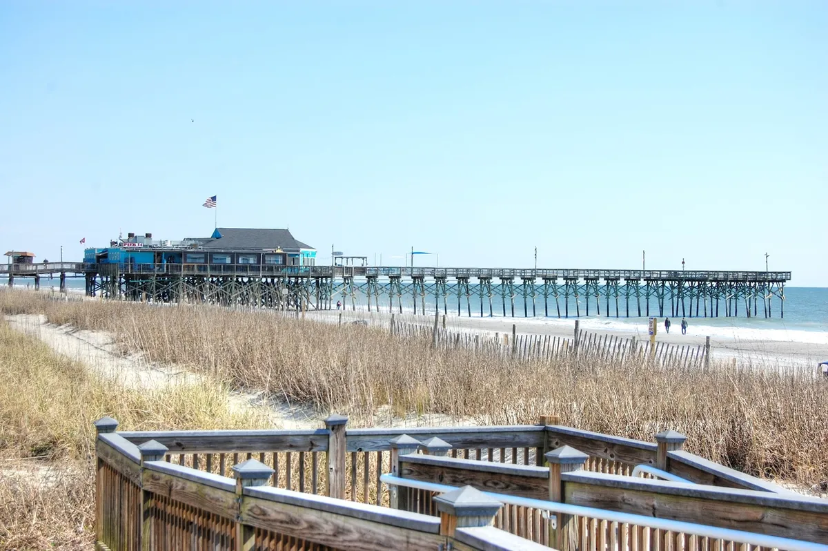 A picturesque view of a classic pier stretching out over the ocean, with soft sand dunes and a wooden boardwalk in the foreground. The tranquil water and open sky evoke relaxation and play, ideal for sandcastles or simply enjoying the sun.