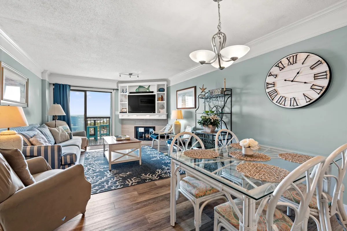 A spacious, open-concept living and dining area featuring comfortable seating, a fireplace, a large clock, and sweeping ocean views visible through a wide window. The dining table is set near the window, and the overall ambiance is cozy and inviting, perfect for family gatherings or relaxing evenings.